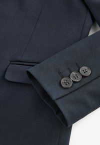 Détail d'une veste de costume bleu foncé montrant un tissu texturé et une manche avec trois boutons noirs et des accents de couture. Conçue de manière ajustée avec un design épuré.