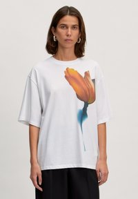 Weißes oversized T-Shirt aus weichem Baumwollstoff mit einem lebhaften orangen Tulpenmotiv auf der Vorderseite. Kurze Ärmel und runder Ausschnitt runden das Design ab.