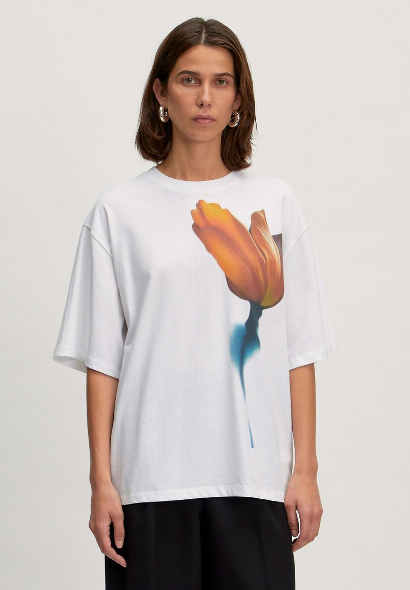 Weißes oversized T-Shirt aus weichem Baumwollstoff mit einem lebhaften orangen Tulpenmotiv auf der Vorderseite. Kurze Ärmel und runder Ausschnitt runden das Design ab.