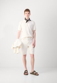 Polo en crème, short en denim beige clair et sandales brunes. Le modèle tient une veste légère assortie. Une écharpe noire à motifs paisley apporte une touche d'accent à la tenue.