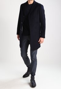 Marinblå ullkappa med slagfickor, tre knappar, i kombination med en svart rundhalsad tröja och slim-fit mörka jeans, ankelstövlar.