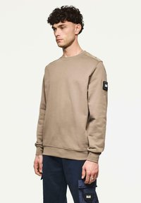 Junger Mann mit lockigem Haar, der einen beigen Sweatshirt und marineblaue Cargo-Hosen trägt, steht vor einem einfarbigen hellen Hintergrund.