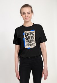Sort T-shirt med et grafisk design af en revnet papirnote med teksten: "UDFORSK! LIDT MERE EVENTYR!" tapet på med blå tape.