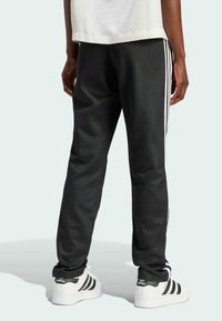 Czarne spodnie sportowe z białymi paskami po bokach, o zwężającym się kroju i miękkim materiale. Noszone z białymi sneakrsami z czarnymi paskami.