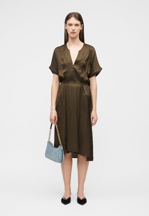 Donna che indossa un vestito midi in seta marrone, tiene una borsa firmata blu con tracolle a catena, in piedi con scarpe nere a punta su uno sfondo bianco.