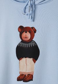Sudadera azul clara con un gráfico de un oso de peluche marrón que lleva un suéter negro con patrones, pantalones beige y botas marrones con detalles en naranja.