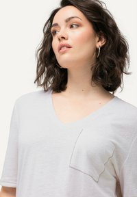 Camiseta blanca de cuello en V hecha de un tejido ligero, con un bolsillo con detalles de costura y mangas cortas enrolladas. Textura suave.