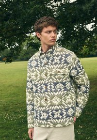 Junger Mann in einer gesteppte Jacke mit geometrischen Blumenmustern in Grün, Weiß und Blau, der im Freien neben einem Baum auf einer Wiese steht.