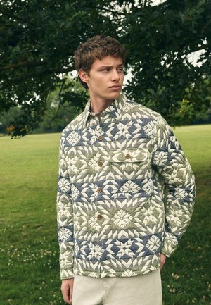 Young man wearing a quilted jacket with geometric floral patterns in green, white, and blue, standing outdoors by a tree in a grassy field.