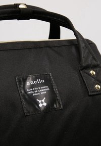 Sac en toile noire avec des détails de fermeture éclair dorée. Présente une forme rectangulaire et une étiquette en tissu avec le nom de la marque et du texte. Poignées incluses.