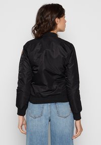 Czarna kurtka bombowa o gładkiej, błyszczącej fakturze, z prążkowanym kołnierzem i mankietami oraz naszywką na lewym rękawie. Do tego jasnoniebieskie jeansy.