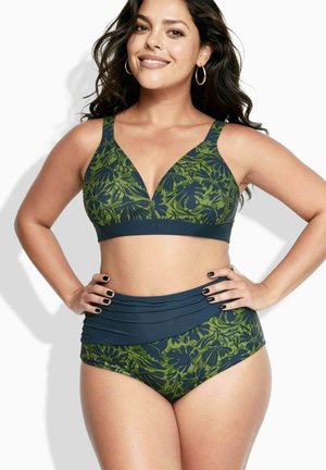 Vrouw met lang donker haar, gekleed in een groene en marineblauwe bikini met hoge taille, glimlachend, met handen in de zij, tegen een witte achtergrond.