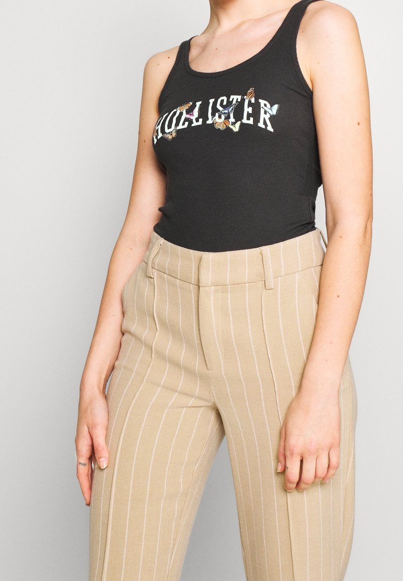 Czarny ribbed tank top z napisem "Hollister" oraz detalami w kształcie motyli, w zestawieniu z beżowymi spodniami o wysokim stanie w paski. Gładka faktura.