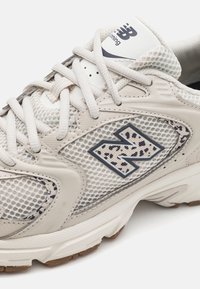 Beige net hardloopschoen met synthetische leren accenten, met een opvallend donkerblauwe "N"-logo en tekstuurdetails aan de zijkant.
