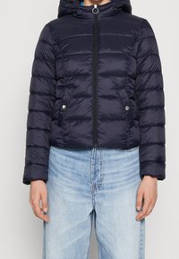 Mörkblå puffjacka med huva, dragkedja framtill och tryckknappar; har sydda paneler och en glänsande finish.