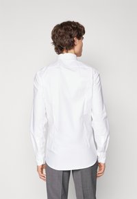 Camicia bianca a maniche lunghe con colletto classico, texture liscia e design della schiena dritto, abbinata a pantaloni grigi a scacchi.