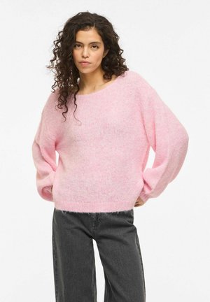 Kobieta o kręconych ciemnych włosach, ubrana w luźny różowy sweter i czarne dżinsy, stojąca z rękami na biodrach na białym tle.