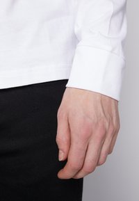 Cămașă albă cu mâneci lungi, cu manșete rulate, asociată cu pantaloni negri. Materialul pare neted, cu cusături vizibile pe margine.