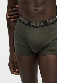 Αρσενικός κορμός που φοράει εσώρουχα Puma σε ελιά πράσινο με μαύρη ζώνη και λογότυπο Puma, το χέρι ακουμπά στον αντίθετο βραχίονα σε λευκό φόντο.