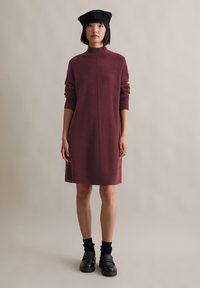 Frau trägt ein burgunderfarbenes Langarm-Strickkleid, eine schwarze Baskenmütze, schwarze Socken und schwarze klobige Loafers und steht vor einem schlichten hellen Hintergrund.