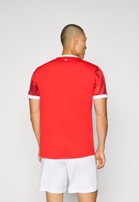 Maglia sportiva rossa a maniche corte, con dettagli bianchi sul colletto e sulle maniche, caratterizzata da un motivo astratto sulle maniche.