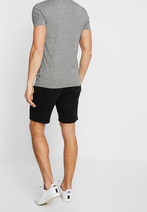Homme debout de dos, portant un t-shirt gris à manches courtes, un short noir et des baskets blanches Adidas sur un fond uni.