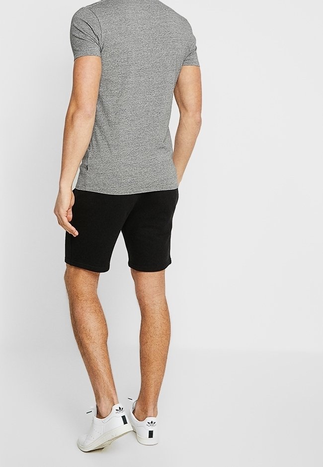 Homme debout de dos, portant un t-shirt gris à manches courtes, un short noir et des baskets blanches Adidas sur un fond uni.