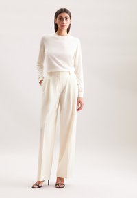 Maglione crema con maniche lunghe abbinato a pantaloni bianchi a vita alta e a gamba larga. Presenta delle pieghe e sandali neri con tacco.