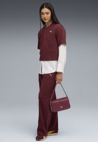 Polo shirt corto bordeaux sopra una camicia a maniche lunghe bianca, abbinato a pantaloni bordeaux e a una piccola borsa a tracolla bordeaux. Presenta il logo Puma.