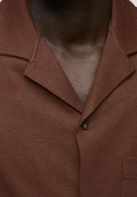 Primer plano de una persona que lleva una camisa marrón con cuello abotonado en el pecho, mostrando la textura de la tela y parte del escote.