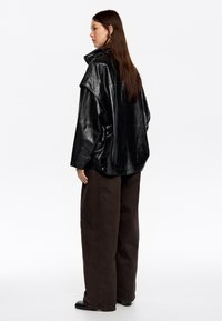 Mujer con cabello largo castaño, vistiendo una chaqueta de cuero negra oversized, pantalones anchos marrones y zapatos negros, de perfil izquierdo sobre un fondo blanco.