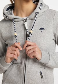 Sudadera gris con cierre y cordón a rayas, detalles de madera y un logo de paraguas bordado en el pecho. Tela suave y texturizada.