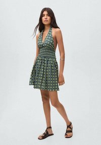 Abito verde con scollo all'americana, corpino arricciato e motivo floreale. La gonna è ampia e abbinata a sandali neri con cinturini.