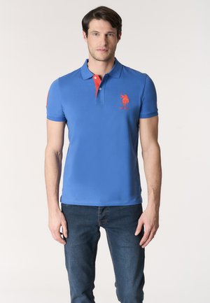 Uomo con polo blu con logo rosso e jeans scuri, in piedi davanti a uno sfondo chiaro uniforme.
