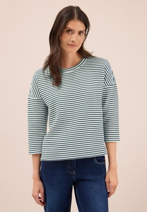 Femme aux longs cheveux bruns portant un haut à manches longues blanc et noir à rayures et un jean bleu foncé, se tenant devant un fond beige.