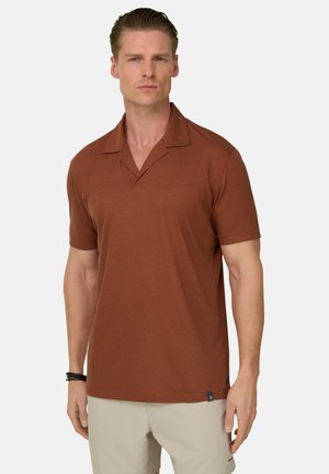 Uomo con capelli corti che indossa una camicia marrone a maniche corte senza colletto e pantaloni beige, in piedi davanti a uno sfondo bianco uniforme.