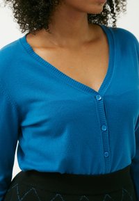 Cardigan en tricot bleu vif avec un col en V, orné de trois boutons bleus et d'une finition côtelée au col et à l'ourlet. Jupe noire visible en dessous.