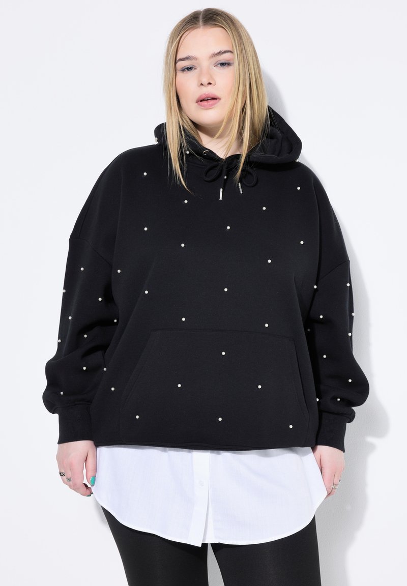 Sudadera con capucha negra de corte holgado, adornada con pequeñas perlas blancas, con un bolsillo frontal y superpuesta a una camisa blanca.