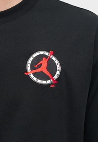 Μαύρο ύφασμα με κόκκινη σιλουέτα παίκτη μπάσκετ που πηδάει με μια μπάλα μέσα σε έναν λευκό κύκλο με το κείμενο "Air Jordan Flight Club".