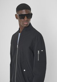 Giubbotto bomber nero con zip frontale, tasche laterali e una piccola tasca con zip sulla manica. Presenta un tessuto testurizzato e un design aderente.