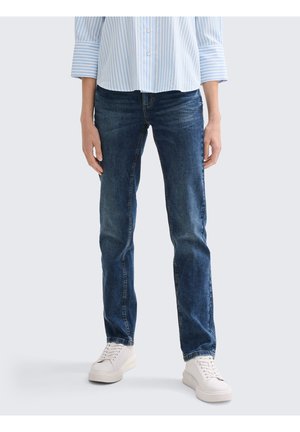 Jean en denim bleu foncé avec une coupe droite, présentant des coutures visibles et une finition soignée, associés à des baskets blanches.