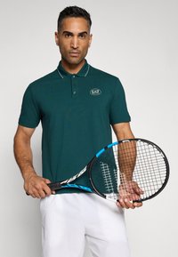 Temno turkizna polo majica z rebrastim ovratnikom in belimi detajli. Ima vezen logotip. Model drži črno-modro teniško lopar.
