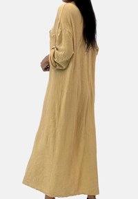 Langes, locker sitzendes Kleid aus strukturiertem senffarbenem Stoff, mit aufgerollten Ärmeln und Seitentasche, bis zur Mitte der Waden reichend.