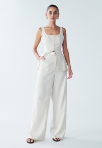 Hellbeiges, ärmelloses Oberteil mit quadratischem Ausschnitt und Brusttasche, kombiniert mit einer weit geschnittenen Hose aus dem gleichen Stoff. Minimalistisches Design, glatte Textur.