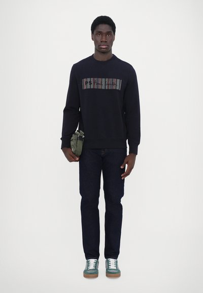Sweatshirt marine avec des rayures horizontales multicolores et du texte, jean foncé, et baskets teal avec des lacets blancs, tenant un petit sac vert.