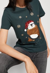 Camiseta de algodón verde oscuro con un gráfico de pingüino, bufanda marrón brillante, gorro rojo con lentejuelas y acentos de copo de nieve dorados.