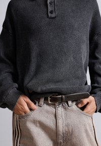 Tummanharmaa ribbikuosis pitkähiha-henley-paita, jossa on kolme nappia ja musta vyö. Kevyesti teksturoitu haalistunut beige farkkuhousut, joissa on sivuraidat.