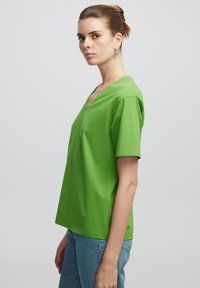 T-shirt verde in cotone a V con maniche corte, caratterizzato da una vestibilità rilassata e una texture liscia; indossato con jeans in denim blu.