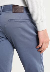 Blå bomullshängselbyxor med en slät textur, som har en brun läderetikett på midjebandet och en bakficka. Slim fit-design.