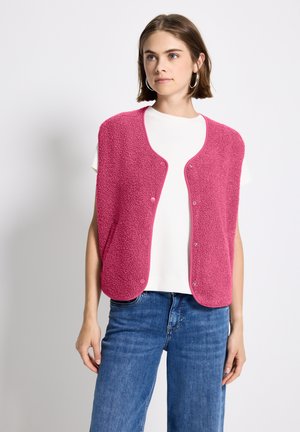 Fuzzy pink vest with snap buttons, rounded edges, and a soft texture, worn over a white top, paired with blue wide-leg jeans.